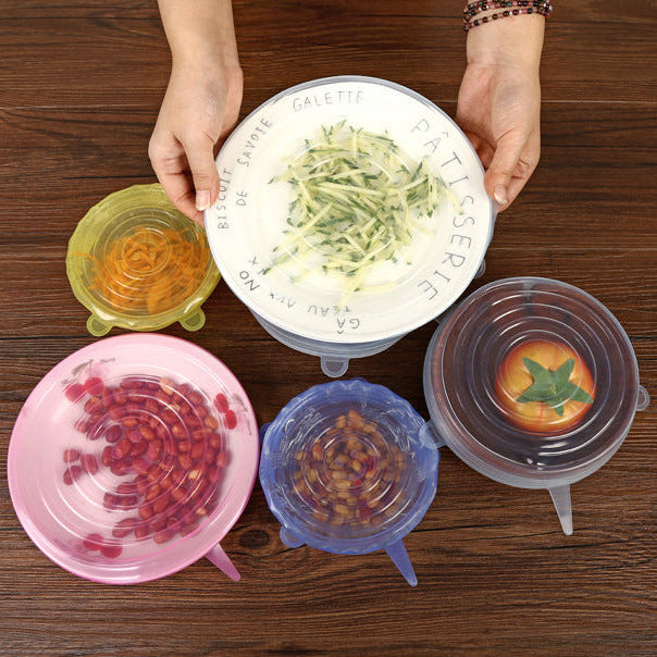 A set of silicone stretchable food storage lids in pink, blue, yellow, and clear, shown with various food items on a dark wooden table. A person's hands are holding a white plate with shredded vegetables to demonstrate the clear lid's use.